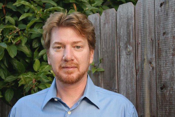 A man with reddish-brown hair and a beard, wearing a blue collared shirt, stands outdoors in front of a wooden fence and gree