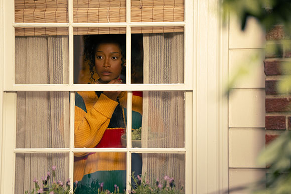 A young Black woman with curly hair looks out a window, resting her chin on her hand. She wears a colorful striped sweater.