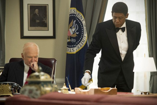 A man in a tuxedo leans over a table to serve a seated man in a dark suit.