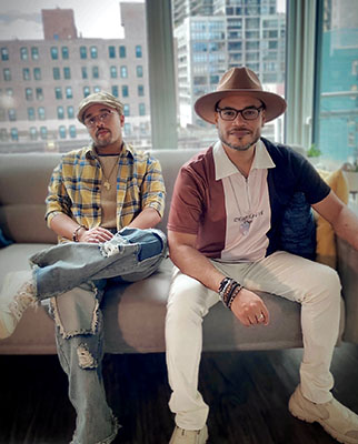 Two men sit on a light-colored sofa in an urban setting with city buildings visible through a large window.