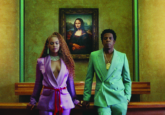 A woman in a purple suit and a man in a teal suit stand before a framed Mona Lisa painting.