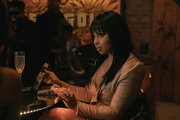 A woman with dark hair and bangs wears a sparkling rose gold dress in a dark lounge setting.