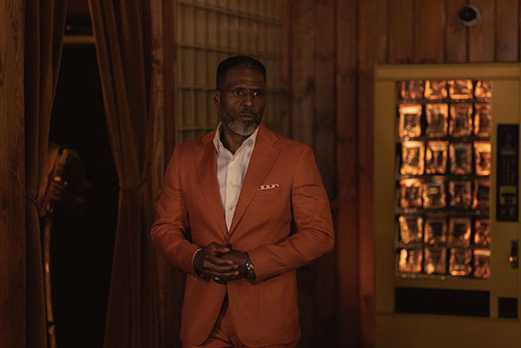 A man with a graying beard wearing a bright orange suit stands in a wood-paneled room.