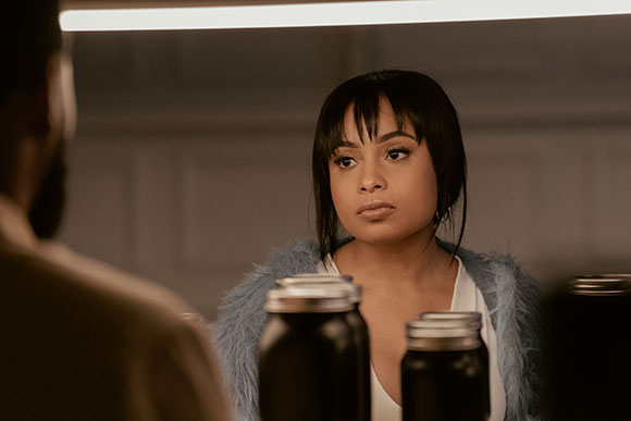 A woman with dark hair and bangs looks toward a man in a blurred foreground.