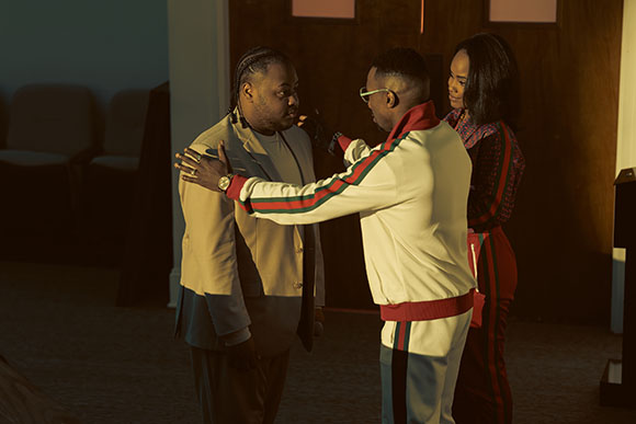 A man in a white tracksuit adjusts the jacket of a man in a light-colored suit while a woman watches.