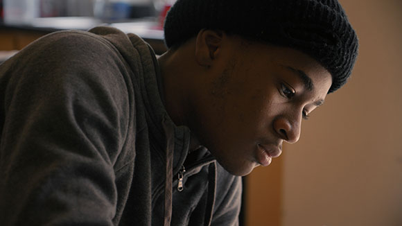 A young man wearing a black beanie and grey hoodie looks downward with a focused expression.