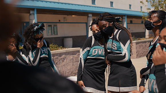 Cheerleaders in black and teal uniforms wearing black face masks stand outdoors near a building.