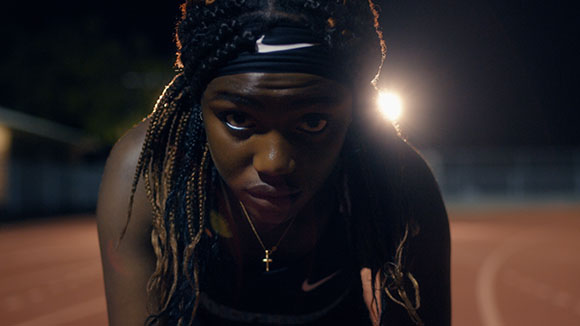 A young woman with long braids and a black headband looks forward at night on a running track.