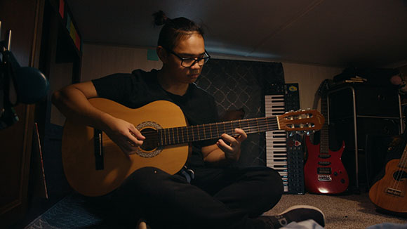 A person wearing glasses sits on the floor playing an acoustic guitar in a dark room with a keyboard behind them.