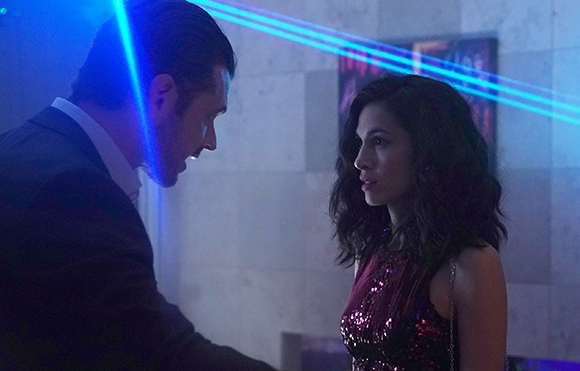 A man in a suit and a woman in a sequined dress look at each other under blue laser beams.