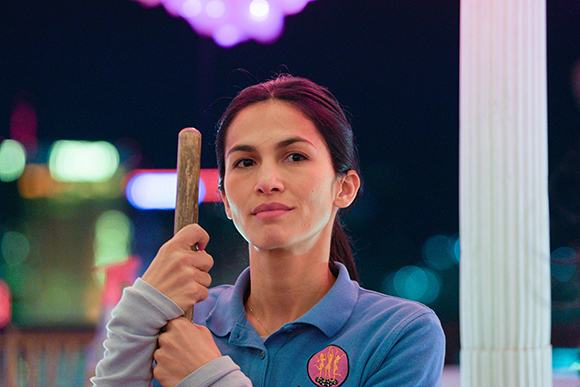 A woman with dark hair pulled back holds a wooden stick against a blurred night background.