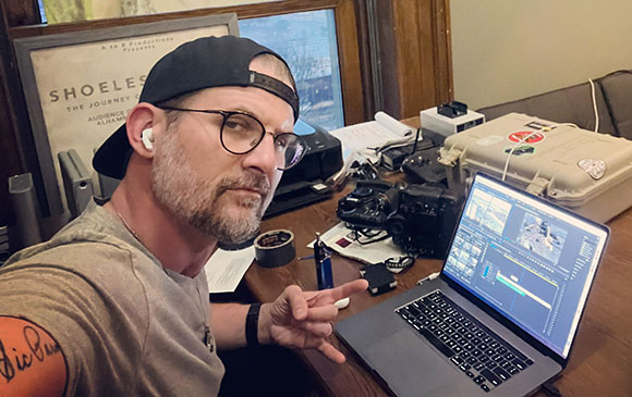A man in a baseball cap and glasses takes a selfie in a room with a laptop and camera gear.