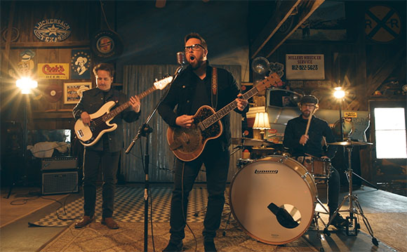 A band performs in a rustic room with a singer, a bassist, and a drummer.
