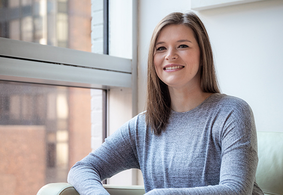 A smiling woman with long brown hair wearing a grey long-sleeved shirt, sitting near a window.