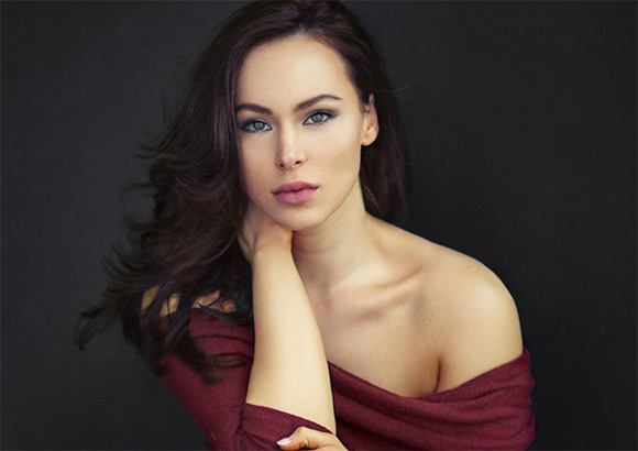 A woman with long dark hair and green eyes looking into the camera, wearing an off-the-shoulder maroon top.