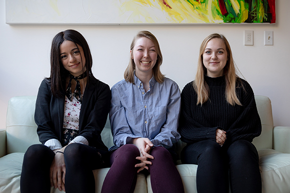 Three young women sitting side-by-side on a light-colored couch against a white wall.