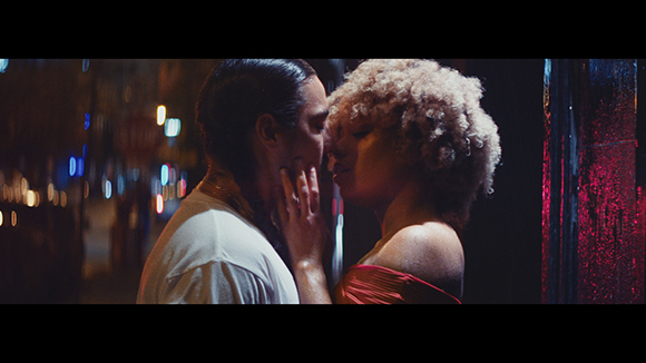 A man with dark hair and a woman with blonde curly hair lean in close to each other at night.