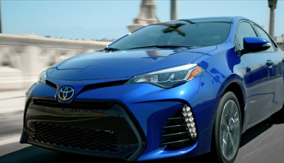 A blue Toyota sedan driving on a paved road under a clear blue sky.