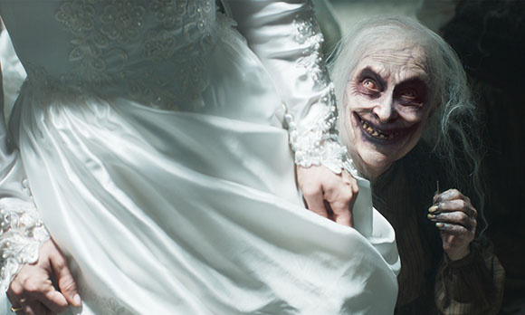 A person in a white gown stands beside a pale, grinning figure with dark eyes and messy hair.