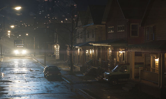 A dimly lit residential street at night with wet pavement and houses with glowing windows.