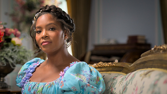 A woman with braided hair wearing a light blue floral puff-sleeve dress sits on an ornate sofa.