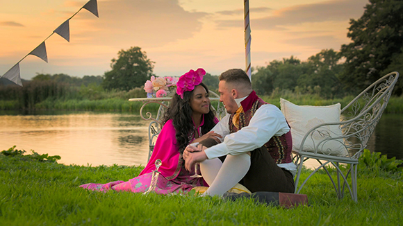 A man and woman sit on a grassy riverbank at sunset, looking at each other near a white metal bench.