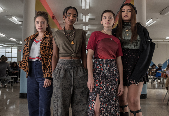 Four teenage girls in diverse casual outfits standing together in a school hallway.
