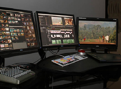 Three computer monitors showing video editing software, a mixing console, and a control surface on a desk.