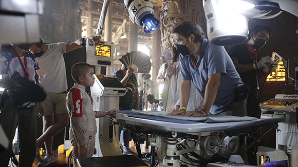 Film crew members work around a young boy sitting near medical equipment on a movie set.