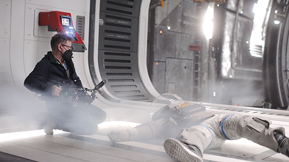 A camera operator kneels with a professional camera, filming a person lying on a foggy sci-fi set floor.