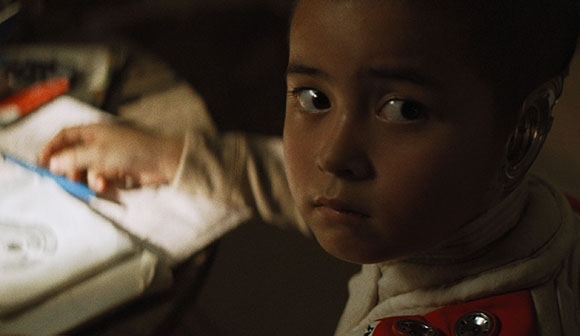 A young child with a metallic device on their ear looks toward the camera while writing in a notebook.