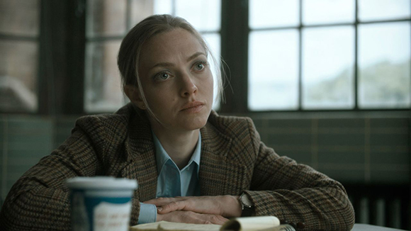 A woman with blonde hair wearing a plaid blazer sits at a table, looking off-camera with a serious expression.
