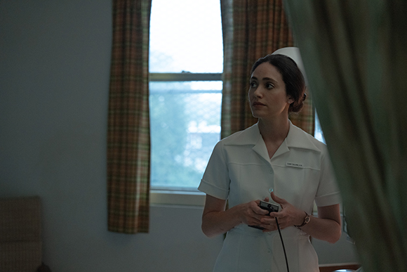A woman in a white nurse's uniform and cap stands in a room near a window and a curtain.