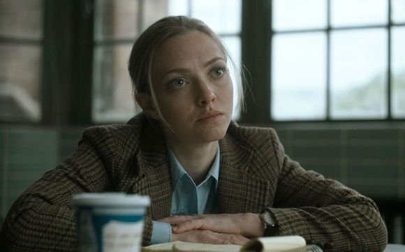 A woman with blonde hair wearing a plaid blazer sits at a table, looking upward with a serious expression.