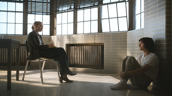 A woman sits in a chair facing another woman sitting on the floor against a tiled wall.