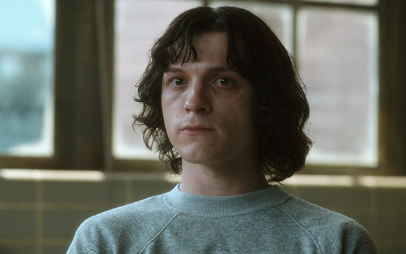 A young man with wavy dark hair and a grey sweatshirt looking forward with a serious expression.