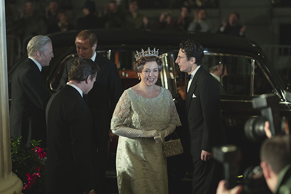 A woman in a light lace gown and tiara smiles while surrounded by men in black formal suits.
