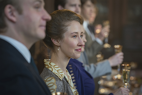 A woman in a patterned blouse smiles while seated at a formal dinner with others in the background.