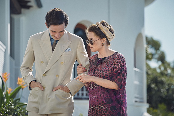 A man in a tan double-breasted suit and a woman in a purple patterned dress walk side by side.
