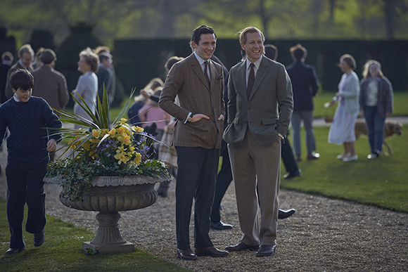Two men in formal blazers and trousers smiling while standing on a gravel path in a garden.