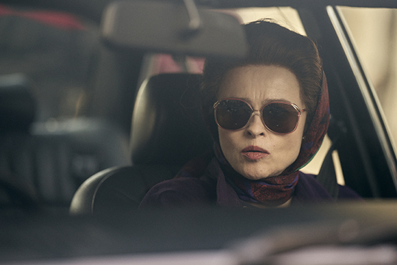 A woman with dark hair, sunglasses, and a patterned headscarf sits in the driver's seat of a car.