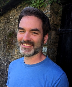 A smiling man with dark hair and a salt-and-pepper beard wearing a blue t-shirt against a stone wall.