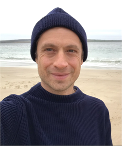 A man wearing a navy blue beanie and ribbed sweater smiles in front of a beach and ocean.
