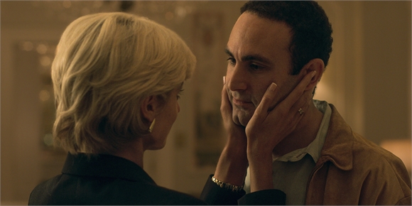 A blonde woman holds a man's face with both hands as they look at each other.