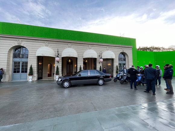 A black sedan parked in front of a classical building facade with a large green screen above it.
