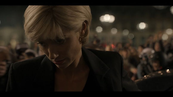 A blonde woman with bangs looks down in a dark, crowded outdoor setting with blurred lights in the background.