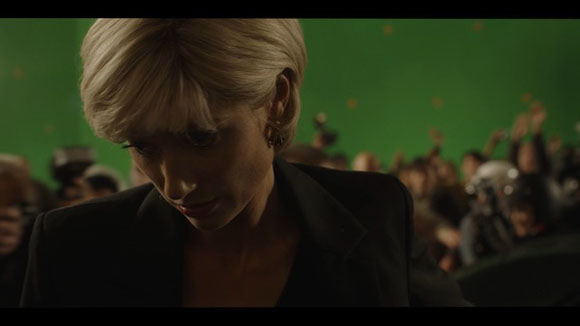 A blonde woman in a dark blazer looks down while a crowd of people stands behind her against a green background.