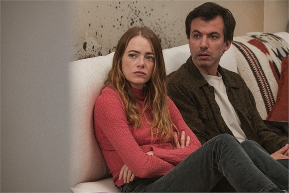 A woman in a pink turtleneck and a man in a patterned shirt sit on a white couch with worried expressions.