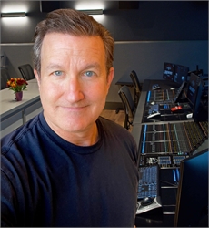 A man in a dark t-shirt smiles in a studio filled with audio mixing consoles and computer monitors.