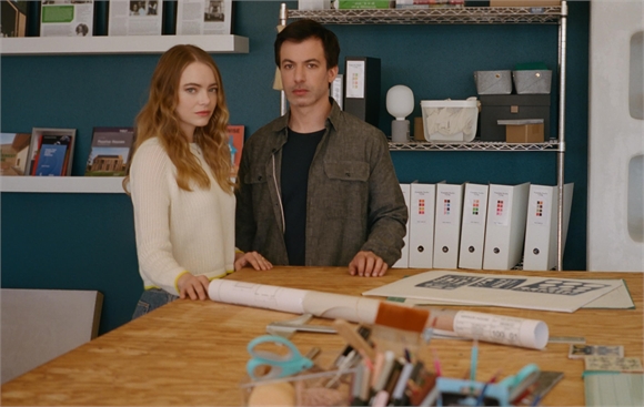 A woman in a white sweater and a man in a dark shirt stand behind a desk with architectural plans.